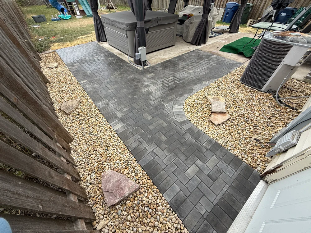 Gray interlocking concrete pavers form a curved patio path in a Saltzman Outdoors hardscaping project in Channahon, Illinois, bordered by gravel and equipment.