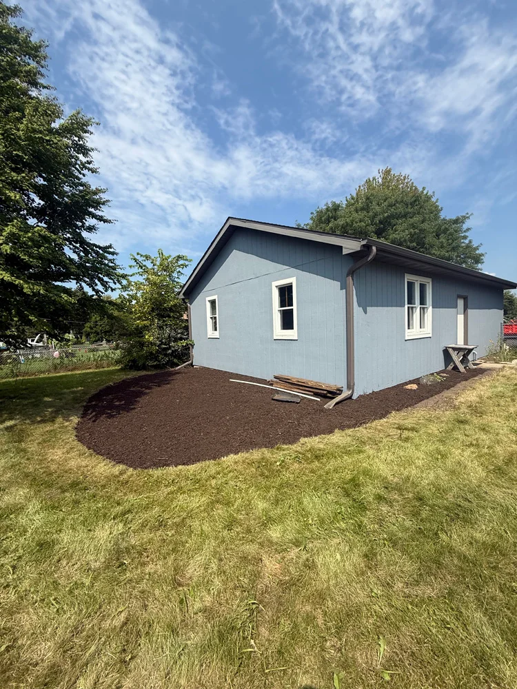 Saltzman Outdoors blue single-story home exterior with landscaped mulch bed Blue single-story house with brown mulch bed in the yard, trees to the left, a clear sky, and a downspout along the side; landscaping renovation context for Saltzman Outdoors