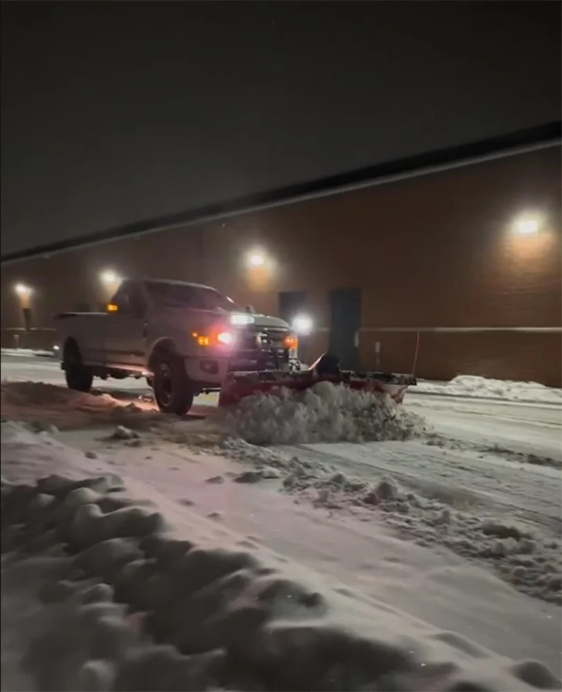 Nighttime snow removal scene with a front-mounted plow on a heavy-duty truck clearing snow from a lot near a brick building, Saltzman Outdoors snow and ice management service.